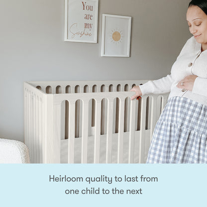A pregnant woman stands beside the Newton Galileo Convertible Crib by Newton Baby in a softly decorated nursery, smiling and touching her belly. Text below reads: Heirloom quality with toddler bed conversion for growing families.