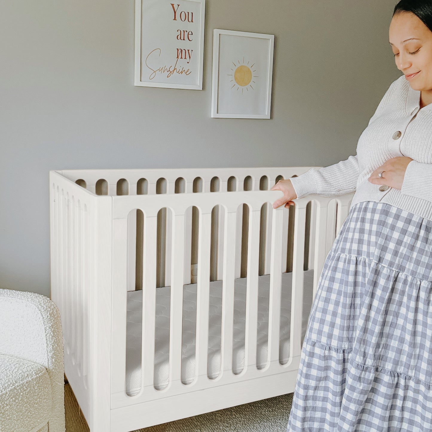 A pregnant woman stands beside the Newton Baby Newton Galileo Convertible Crib in a softly lit nursery, with framed art on the walls—one saying "You are my sunshine," another featuring a sun—gently touching her belly.