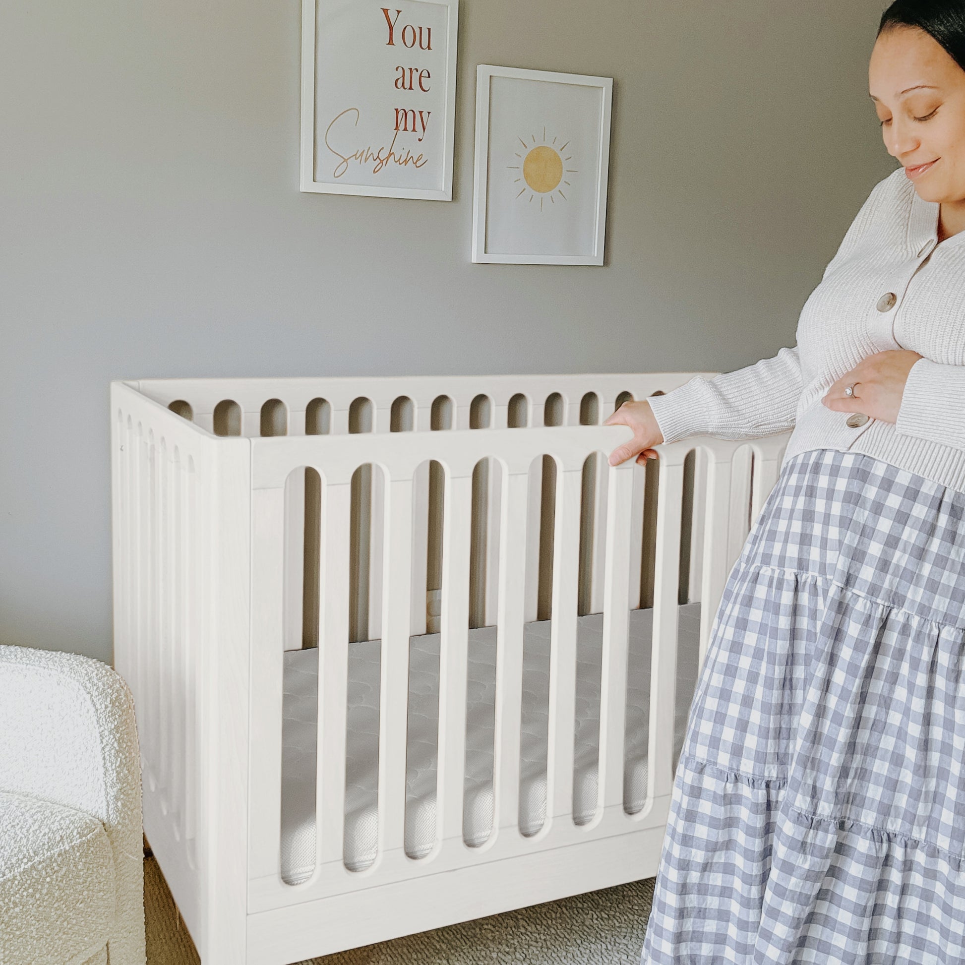 A pregnant woman stands beside the Newton Baby Newton Galileo Convertible Crib in a softly lit nursery, with framed art on the walls—one saying "You are my sunshine," another featuring a sun—gently touching her belly.