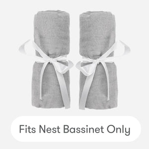 Two gray cotton muslin Nest Bassinet Extra Sheets by Newton Baby, rolled and tied with white ribbons, side by side on a light background. Text below: Fits Nest Bassinet Only—perfect breathable crib sheets for your baby.
