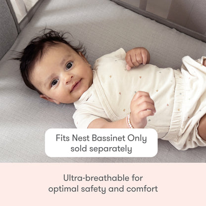 A baby rests in a light-colored bassinet wearing a cream outfit with heart prints. Text: Newton Baby Nest Bassinet Extra Sheets—ultra-breathable, for optimal safety and comfort. Fits Nest Bassinet only (sold separately).