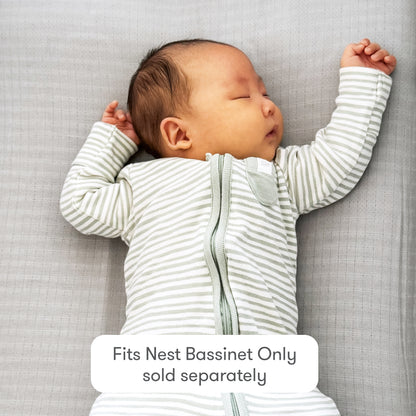 A baby in a gray and white striped onesie sleeps on a light gray surface, arms raised. Text overlay: Newton Baby Nest Bassinet Extra Sheets—cozy cotton muslin, breathable comfort. Fits Nest Bassinet only; bassinet sold separately.