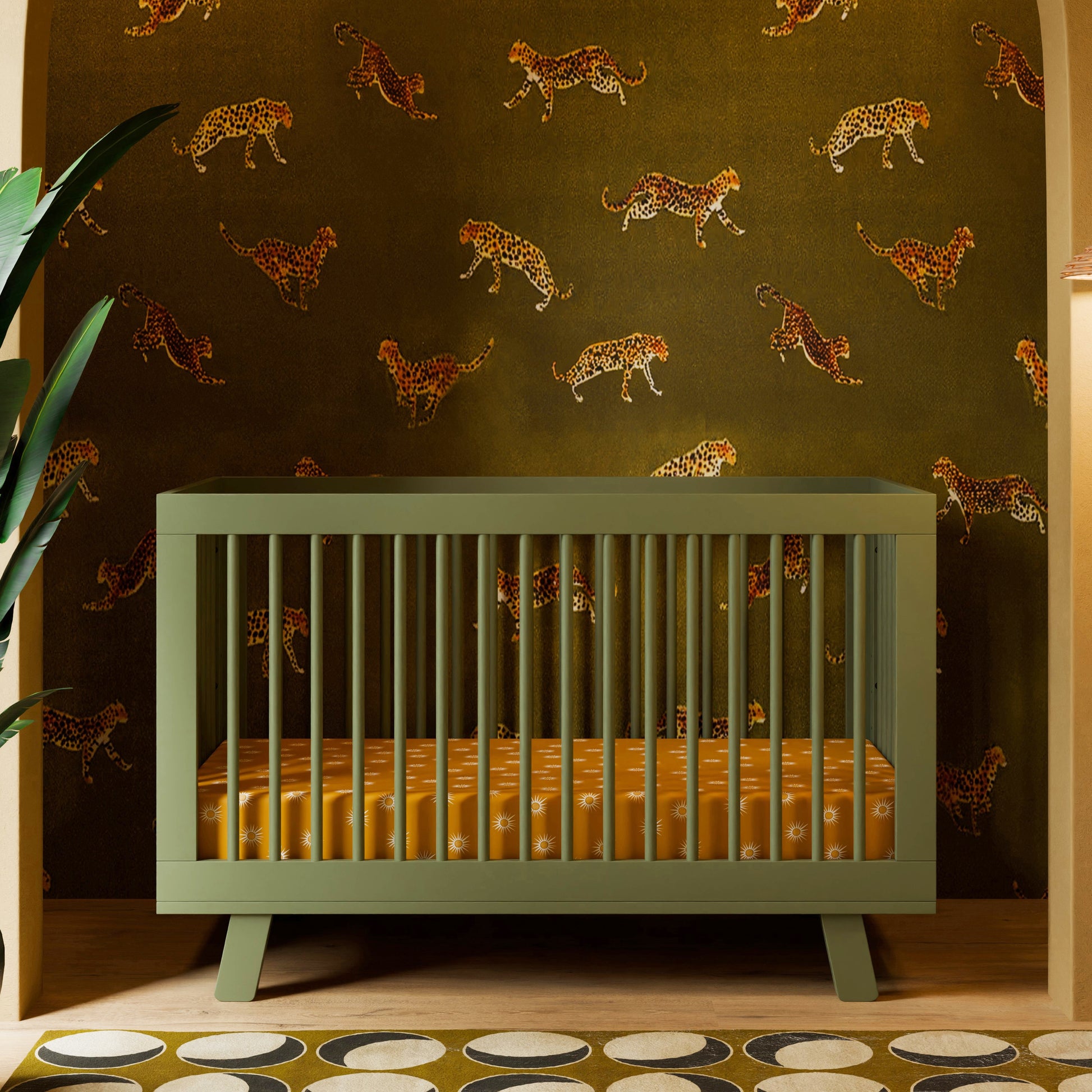 A green Babyletto Hudson Convertible Crib with an orange patterned mattress sits before leaping cheetah wallpaper, accented by a big leaf on the left and a retro circle rug for playful charm.