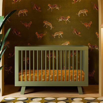 A green Babyletto Hudson Convertible Crib with an orange patterned mattress sits before leaping cheetah wallpaper, accented by a big leaf on the left and a retro circle rug for playful charm.