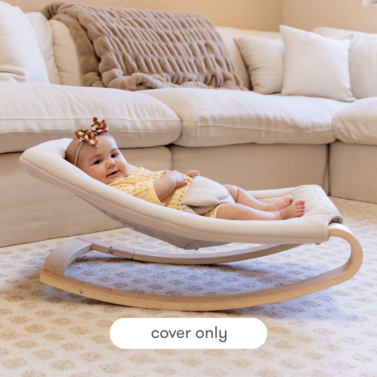 A happy baby dressed in yellow rests on a Newton Baby Rocker fitted with the Baby Rocker Extra Cover in a cozy living room, where a beige sofa and blanket are visible in the background. Text at the bottom reads, cover only.