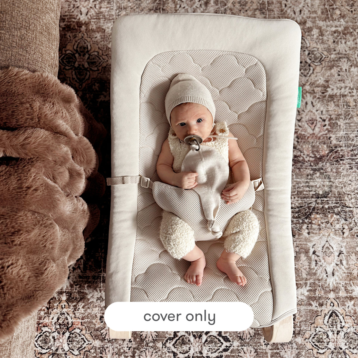 A baby dressed in a beige outfit and hat lounges on a cream-colored Newton Baby Rocker equipped with the Extra Cover on top of a patterned rug. A pacifier clip is attached to the baby’s outfit. The image includes text that states, “cover only.”.