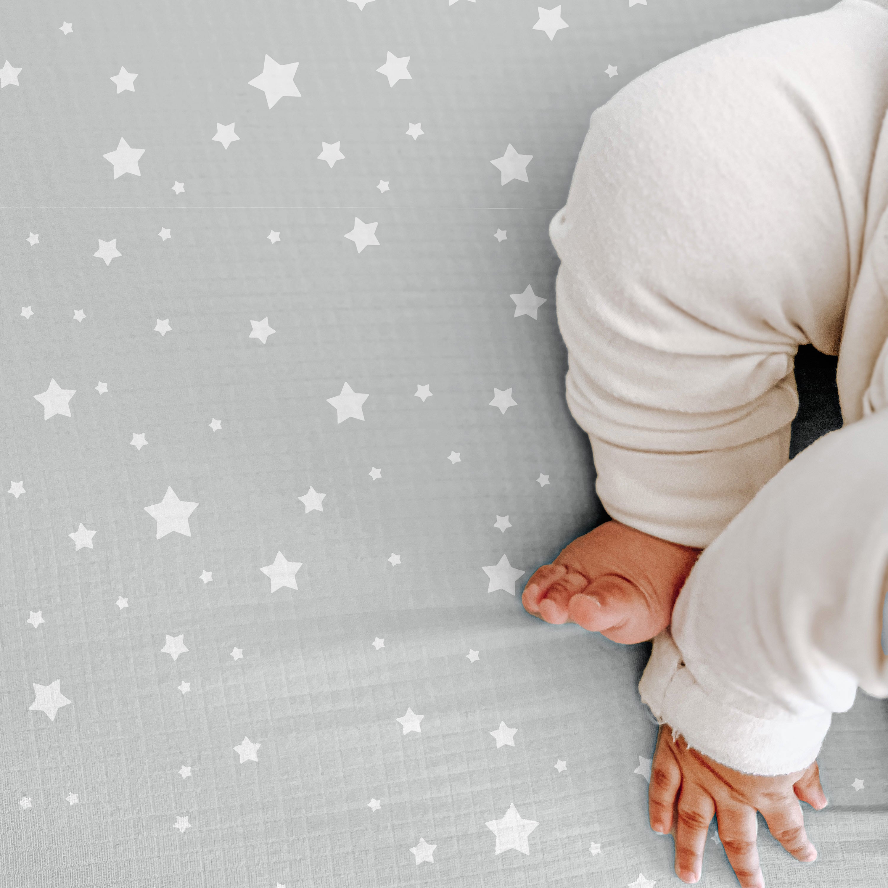 A baby’s legs, feet, and hand rest on a gray blanket with white stars, highlighting the comfort of Newton Baby Breathable, Organic Cotton Sheets (2-pack), crafted for softness and hypoallergenic safety.