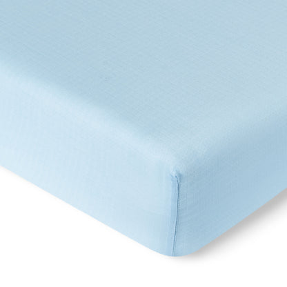 Close-up of the corner of a Newton Baby Breathable, Organic Cotton Sheet (from the 2-pack), in light blue, covering a mattress. The image highlights the smooth, slightly textured fabric and neatly finished seams.