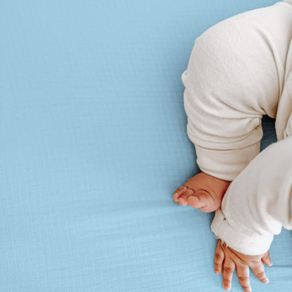 A baby, partially visible, rests on a light blue Newton Baby Breathable, Organic Cotton Sheet (2-pack), known for its soft and hypoallergenic fabric.