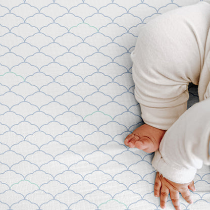 A baby in a white outfit sits on a patterned blanket, showing just their lower half—perfect for highlighting Newton Baby’s Breathable, Organic Cotton Sheets (2-pack).