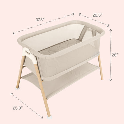 The Newton Baby Essential Bassinet in beige showcases breathable mesh sides, sturdy wooden legs, and a handy lower storage shelf. Measuring 37.8″ long, 20.5″ wide, and 28″ high with legs spaced 25.8″ apart, it is displayed on a soft pale pink background for an elegant touch.