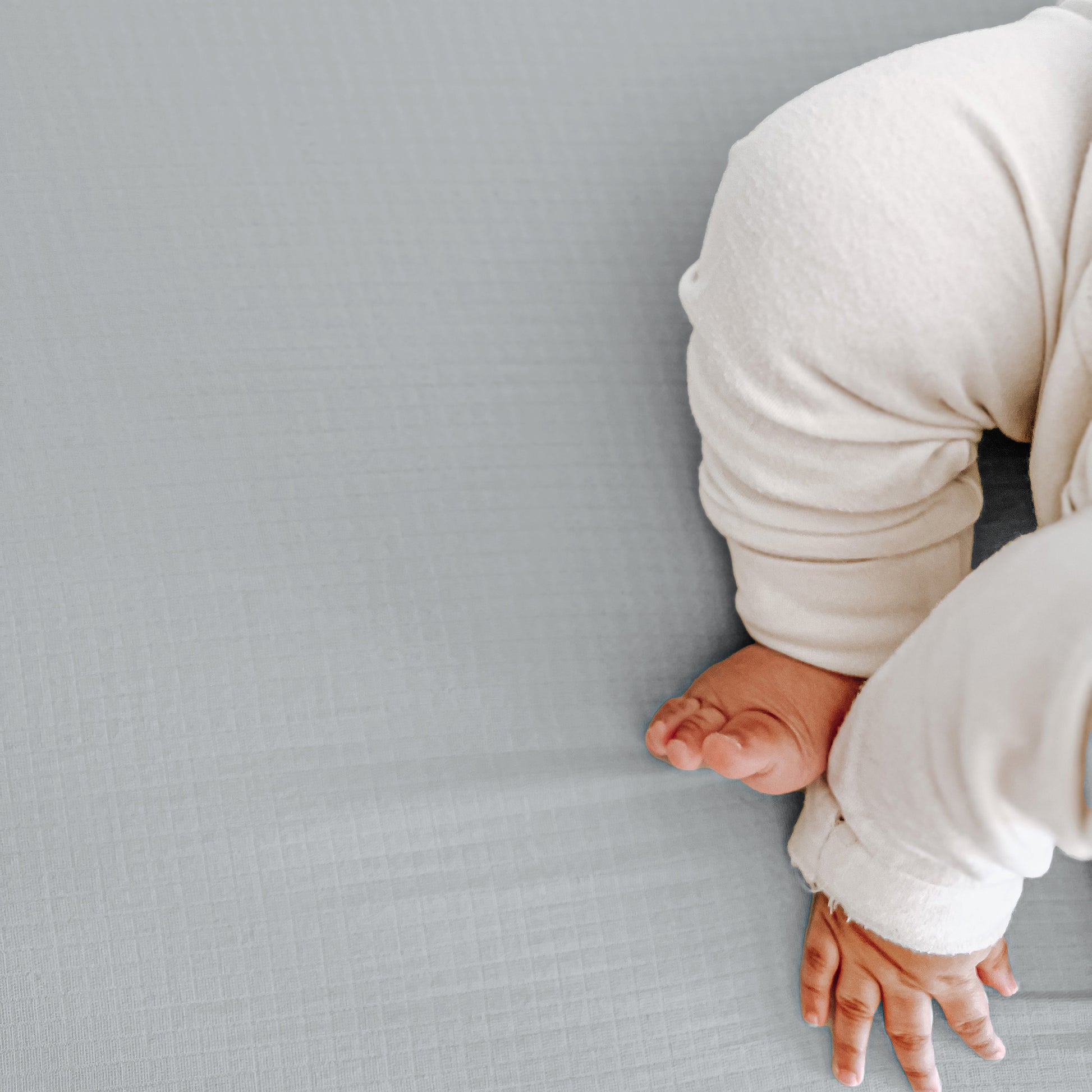 A baby in white pajamas sits on a light gray textured surface, with only the lower half visible and one hand resting on Newton Baby Breathable, Organic Cotton Sheets (2-pack).