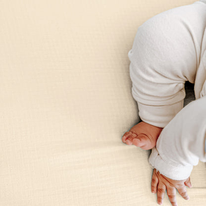 A baby, with only legs and one hand visible, sits on a cream-colored textured surface—likely Newton Baby’s Breathable, Organic Cotton Sheets (2-pack).