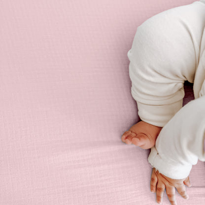 A baby in white relaxes on a soft, pale pink blanket, highlighting Newton Baby’s Breathable, Organic Cotton Sheets (2-pack)—the perfect cozy crib sheets for your little one.