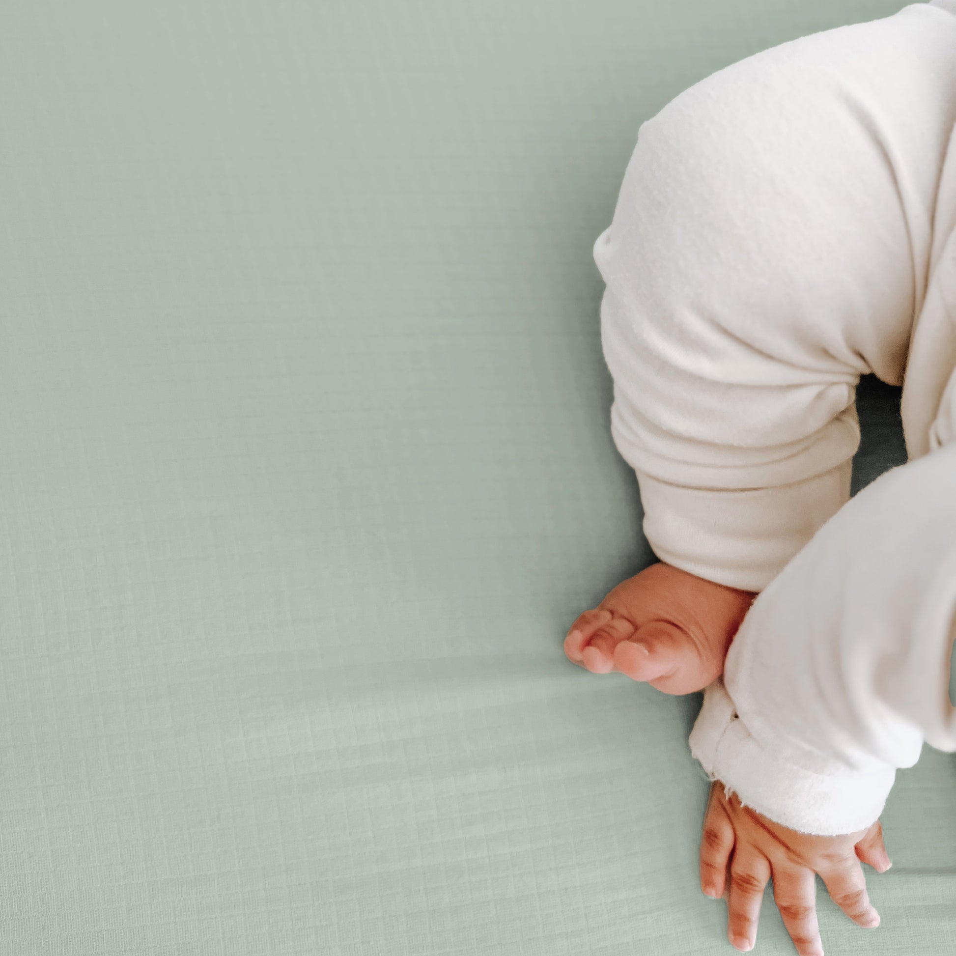 A baby in white pajamas sits on a pale green surface, with just their lower body and hand in frame—perfect for highlighting Newton Baby’s Breathable, Organic Cotton Sheets (2-pack).