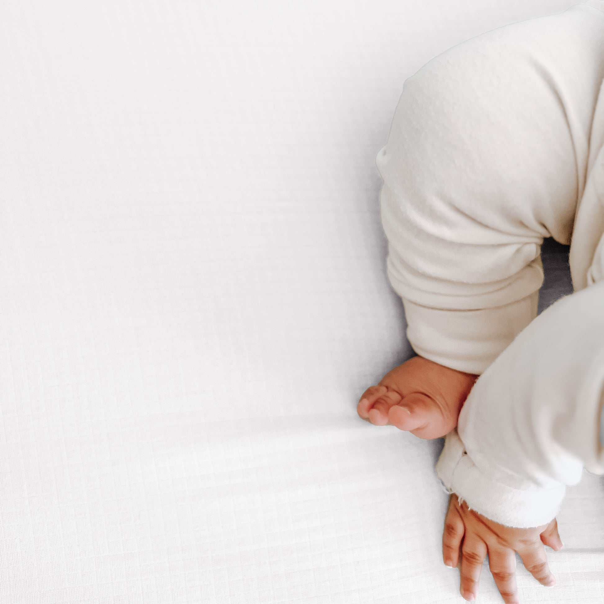 A baby in a white outfit lies on Newton Baby Breathable, Organic Cotton Sheets (2-pack), with only the legs and one hand visible. The scene is gentle and minimalistic.