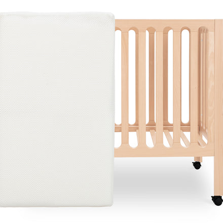 A white crib mattress stands upright in front of a light wood baby crib with vertical slats and two visible wheels on the right legs. The scene is minimal with a plain white background.