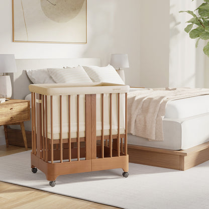 The Newton Baby Newton Nest Convertible Crib with mattress and vertical slats stands beside a modern bed in a bright, cozy bedroom featuring soft bedding, light wood furniture, and a large leafy plant.