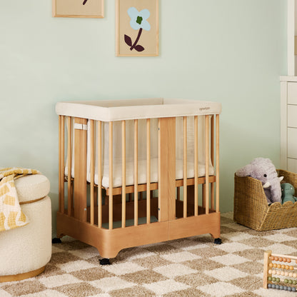 The Newton Baby Newton Nest Convertible Crib with a cream mattress sits on a checkered rug in a softly lit nursery. Nearby are a basket of stuffed animals, a dresser, and flower artwork on the mint-green wall.