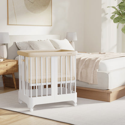 A modern bedroom featuring the Newton Baby Newton Nest Convertible Crib, which is GREENGUARD Gold certified, positioned beside a bed and highlighted by neutral tones, soft bedding, wooden furniture, and natural light streaming through a window.
