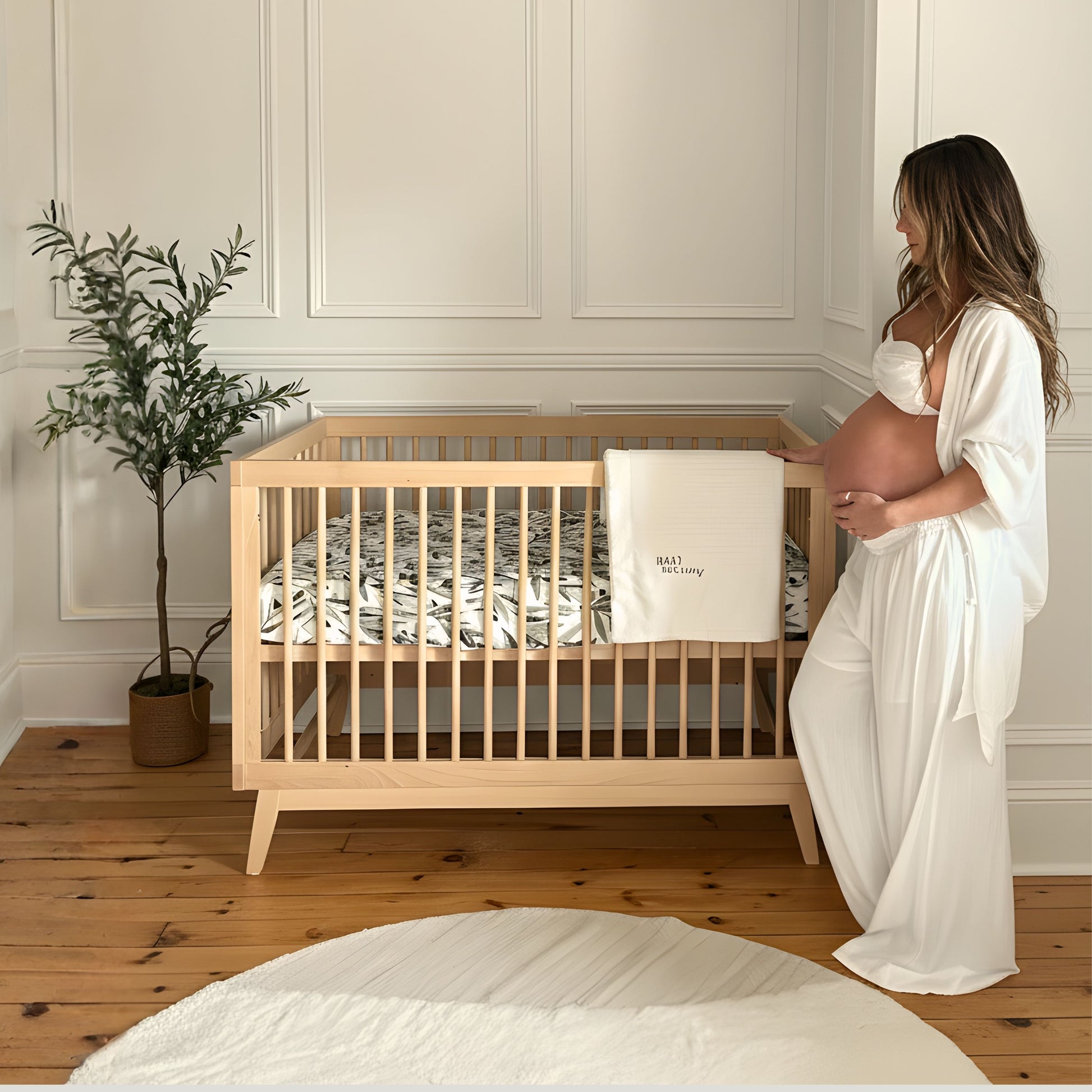 A pregnant woman in white stands beside the dadada baby Soho Convertible Crib with a leafy-patterned Newton crib mattress, in a softly lit, minimalist nursery featuring mid-century accents, a potted plant, and white paneled walls.