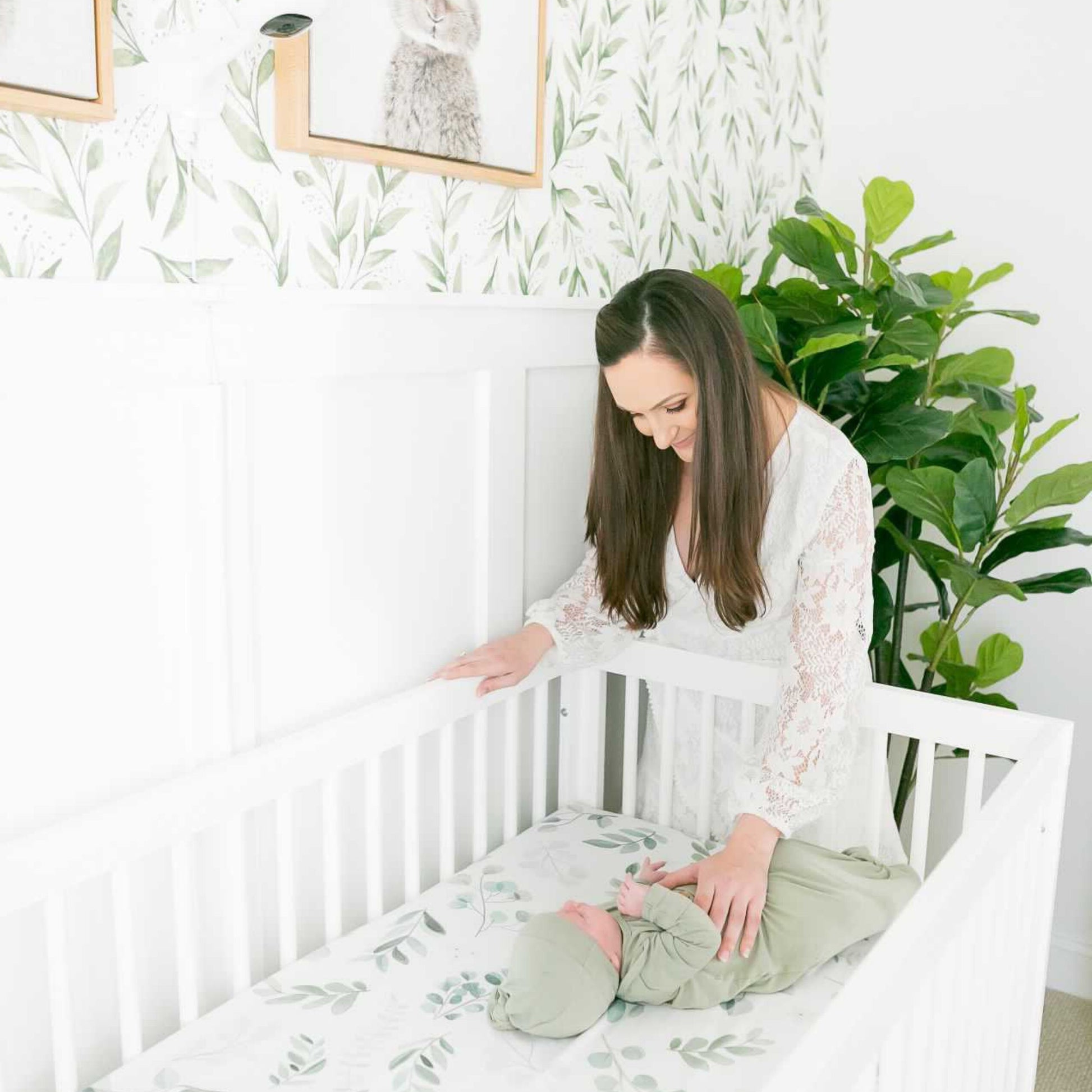 A woman in a white dress stands by the dadada baby Soho Convertible Crib, gently touching a baby in green. The nursery features leafy wallpaper, framed animal art, and a large green plant in the corner.