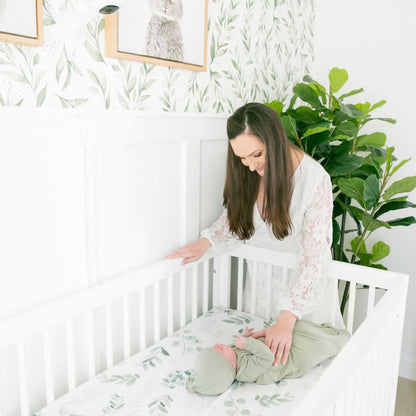 A woman in a white dress stands by the dadada baby Soho Convertible Crib, gently touching a baby in green. The nursery features leafy wallpaper, framed animal art, and a large green plant in the corner.