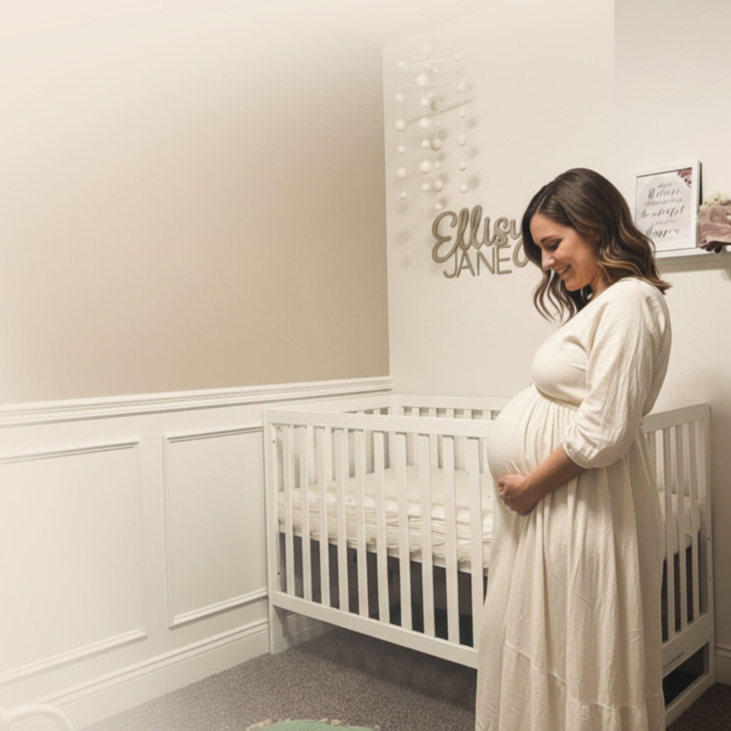 A pregnant woman in a white dress stands beside a dadada baby Austin Convertible Crib in a softly lit nursery, gently holding her belly and smiling. A sign above the crib displays the name Ellie Jane.