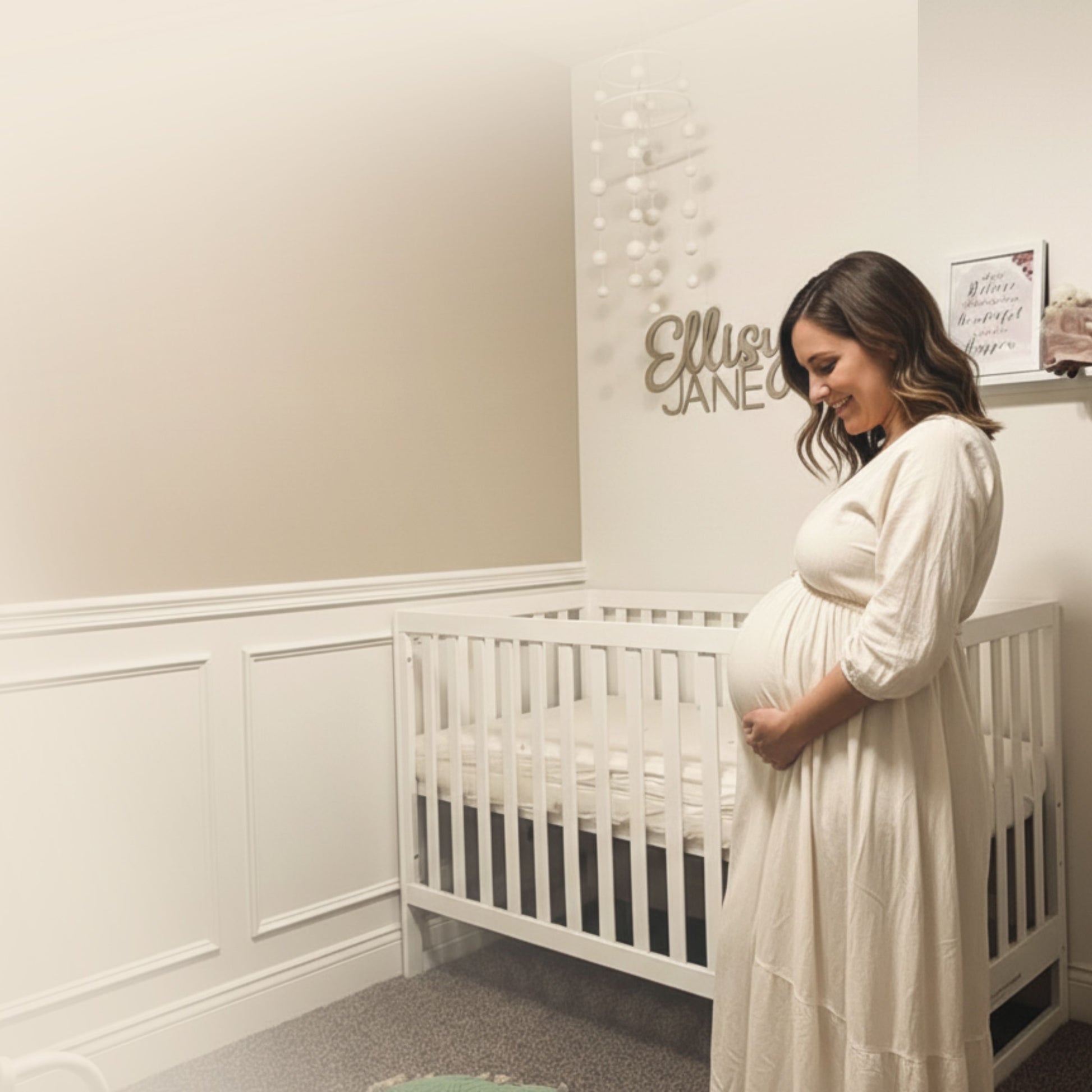 A pregnant woman in a white dress stands beside a dadada baby Austin Convertible Crib in a softly lit nursery, gently holding her belly and smiling. A sign above the crib displays the name Ellie Jane.