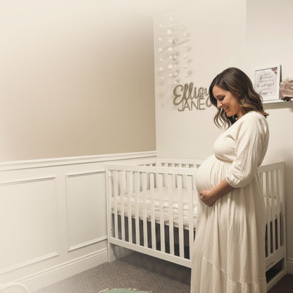 A pregnant woman in a white dress stands beside a dadada baby Austin Convertible Crib in a softly lit nursery, gently holding her belly and smiling. A sign above the crib displays the name Ellie Jane.