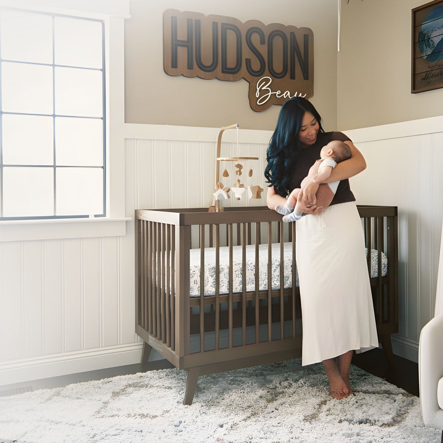 A woman holding a baby stands beside a dadada baby Soho Convertible Crib with a Newton crib mattress in a softly lit, mid-century style nursery. "HUDSON Beau" is above the crib, with a large window, mobile, and plush rug in the room.