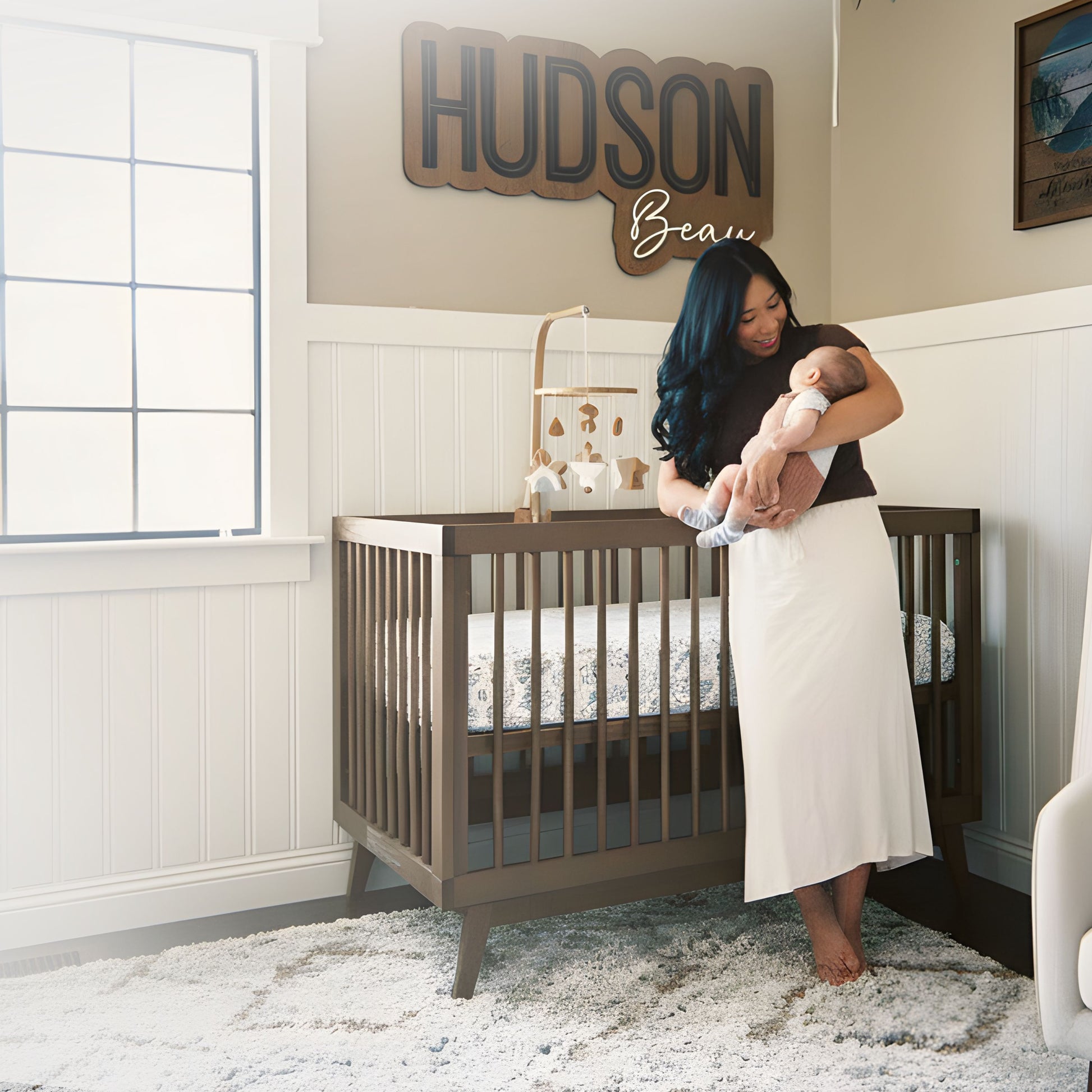 A woman holding a baby stands beside a dadada baby Soho Convertible Crib with a Newton crib mattress in a softly lit, mid-century style nursery. "HUDSON Beau" is above the crib, with a large window, mobile, and plush rug in the room.