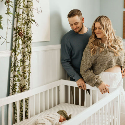 A smiling couple stands by the dadada baby Soho Convertible Crib, gazing at their swaddled baby. The bright nursery has light blue walls and floral curtains, enhancing the cozy space around this modern mid-century style crib.