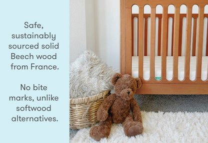 A plush teddy bear sitting beside a wooden crib with text overlay that reads "Safe, sustainably sourced solid Beech wood from France. No bite marks, unlike softwood alternatives."
