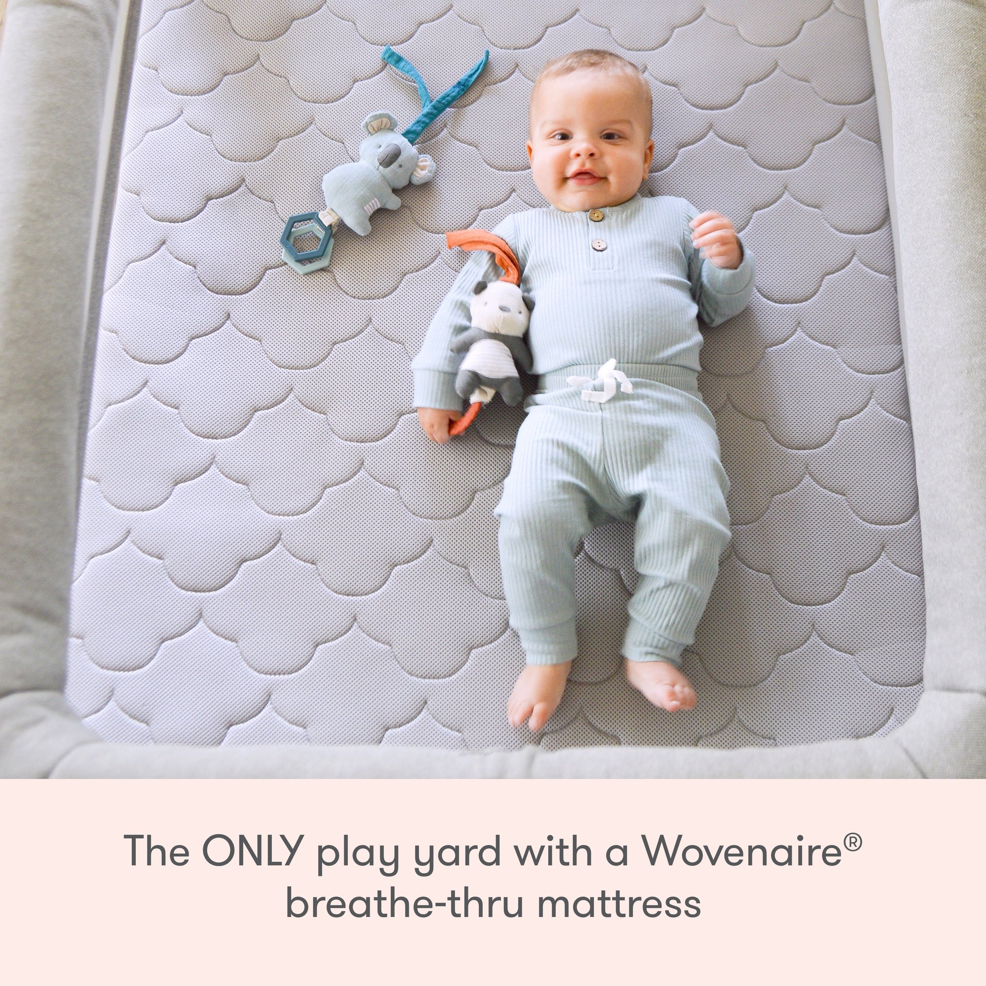 A baby rests on a gray mattress in the Deluxe Travel Crib & Play Yard by Newton Baby, cuddling a stuffed animal. Text below reads: The ONLY travel crib with a Wovenaire® breathe-thru breathable mattress.