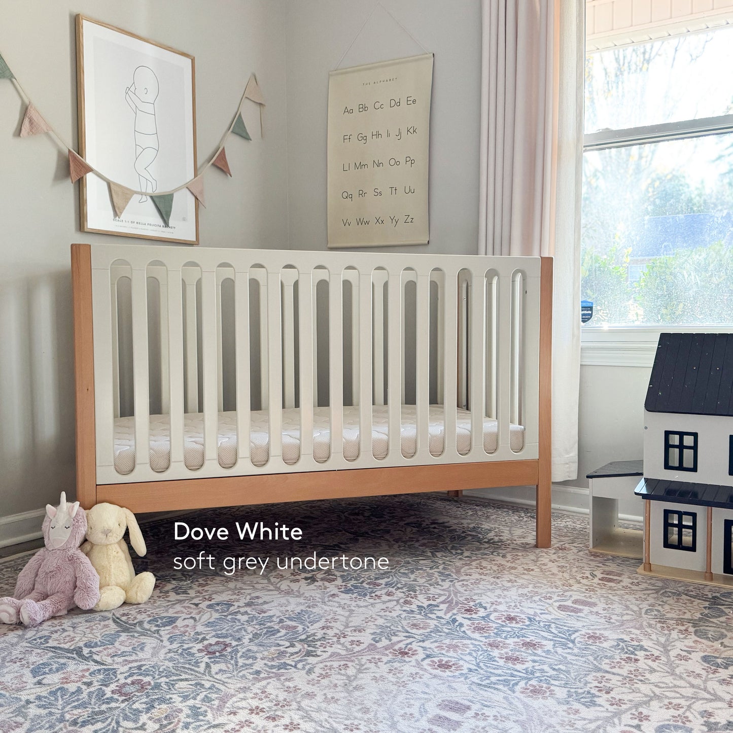 A modern nursery featuring the Newton Baby Newton Galileo Convertible Crib in beech wood and white, a stuffed unicorn and bunny on a patterned rug, dollhouse, alphabet poster, bunting. Text: Dove White soft grey undertone. Shown with toddler bed conversion kit. 999-8420-NDW