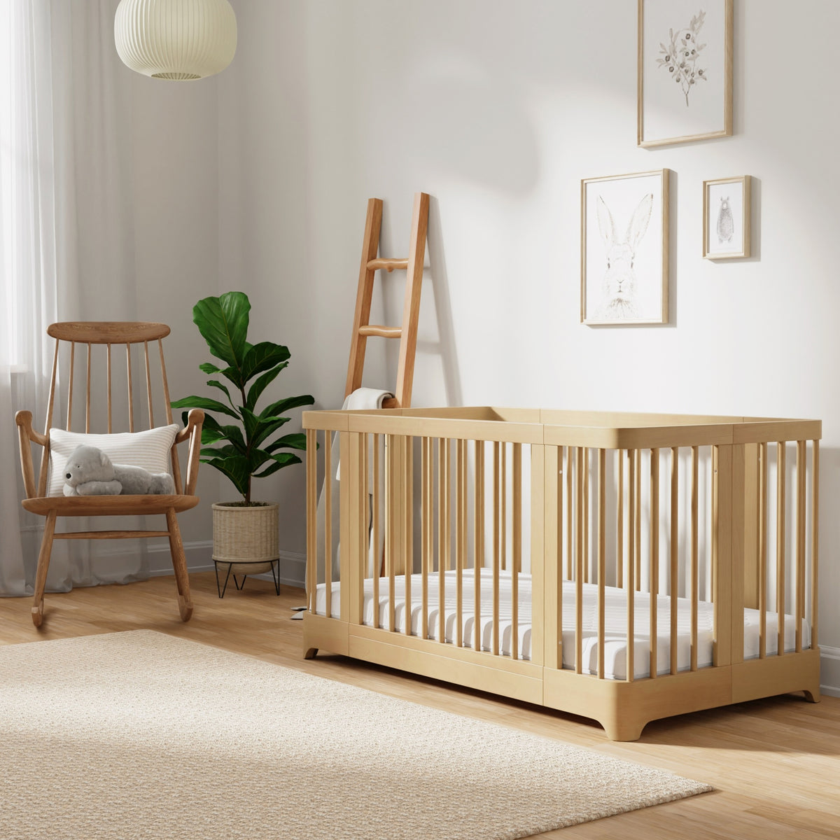 A cozy, modern nursery showcases the Newton Baby Newton Nest Convertible Crib (GREENGUARD Gold certified), a rocking chair with a cat pillow, potted plant, ladder shelf, and framed animal art in gentle natural light.