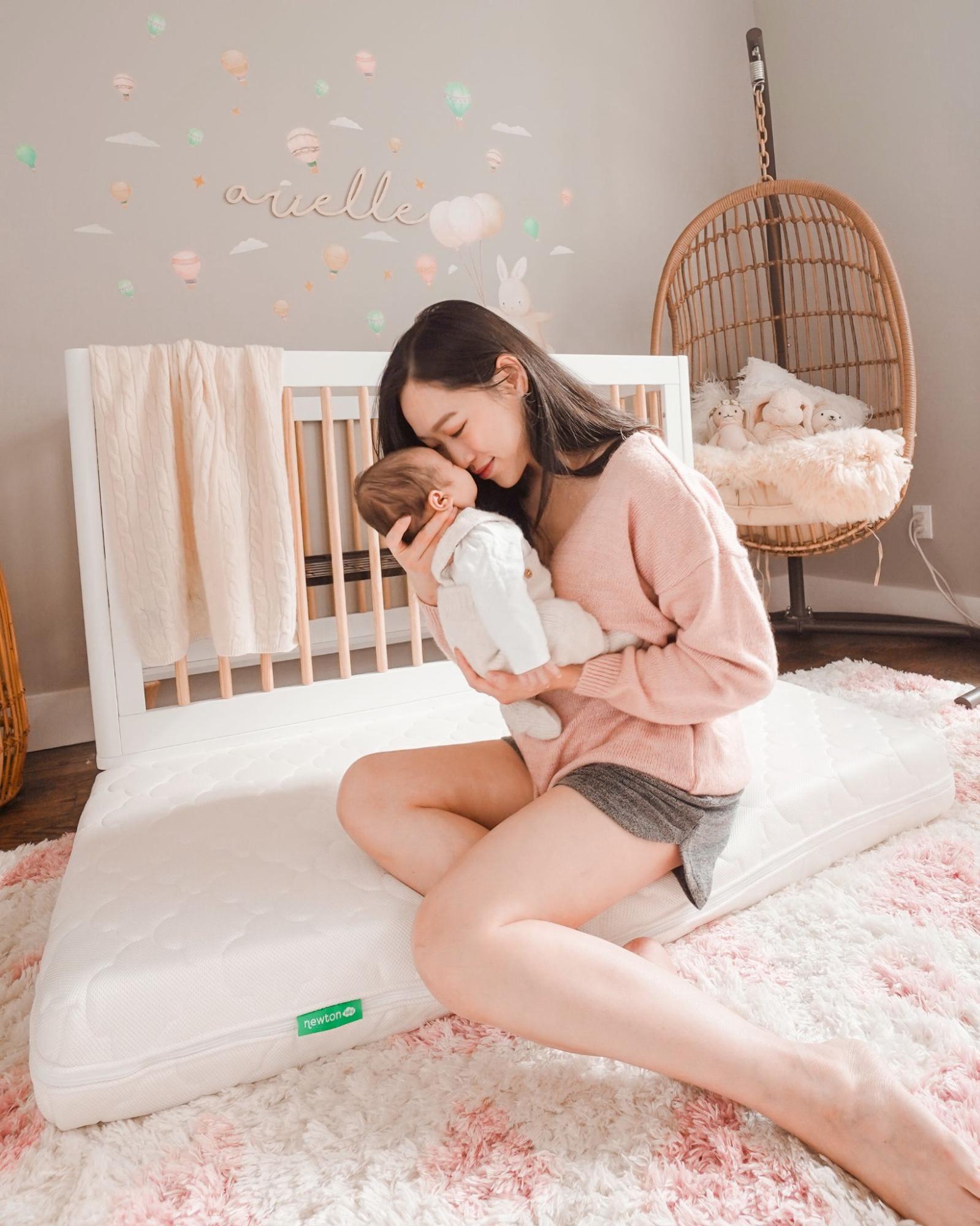 A woman sitting on a mattress in a nursery, gently holding and touching noses with a baby. Behind them is a white crib, wall art, and a hanging chair with stuffed animals. The scene is bright and cozy.