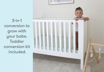 Toddler peeking over the side of a white 3-in-1 convertible crib, with text stating "3-in-1 conversion to grow with your babe. Toddler conversion kit included."