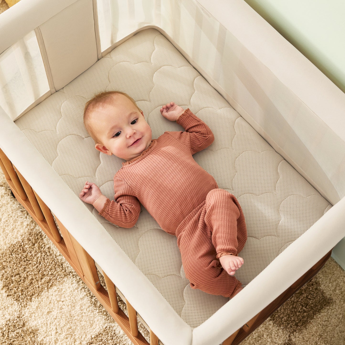 A baby in a rust-colored outfit smiles up while lying in the Newton Nest Convertible Crib by Newton Baby, which includes a mattress and is GREENGUARD Gold certified. The crib rests on a beige and brown patterned rug for added warmth.
