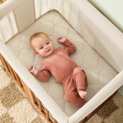 A baby in a rust-colored outfit smiles up while lying in the Newton Nest Convertible Crib by Newton Baby, which includes a mattress and is GREENGUARD Gold certified. The crib rests on a beige and brown patterned rug for added warmth.