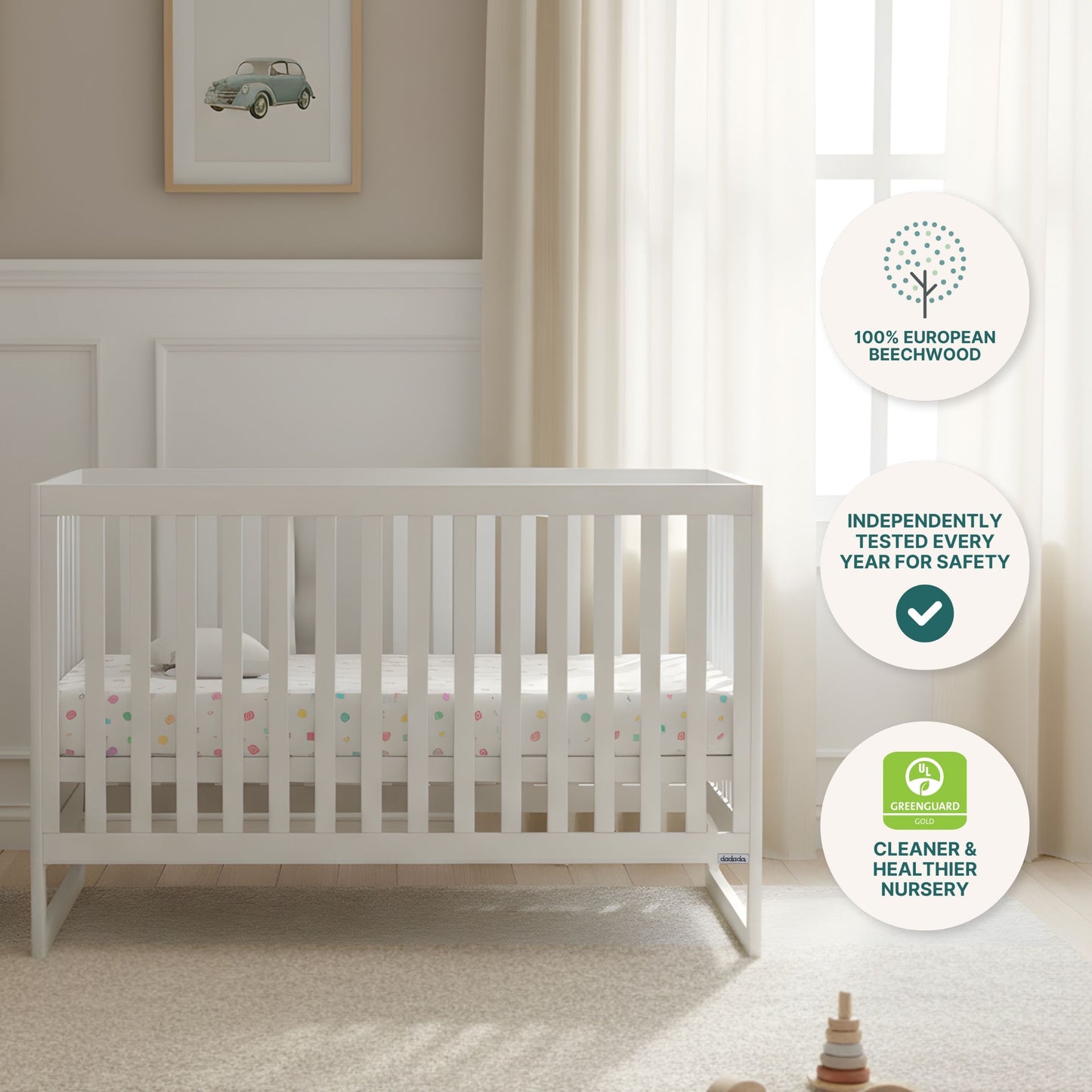 A bright nursery features the dadada baby Austin Convertible Crib in white with a Newton crib mattress, cream curtains, car wall art, and three circular icons for sustainable beechwood, safety testing, and GREENGUARD certification.