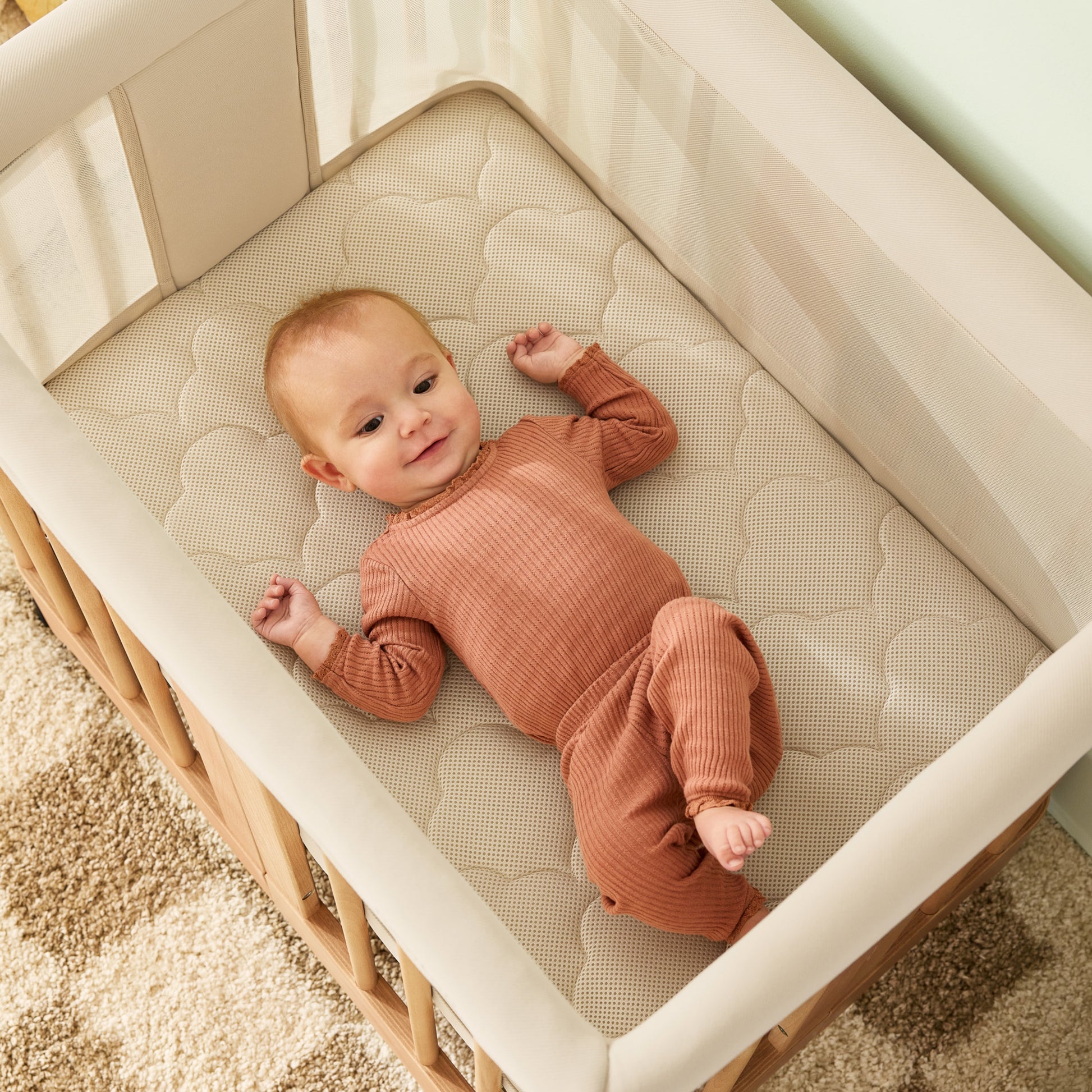 A baby in a rust ribbed outfit smiles while lying on their back in the Newton Baby Newton Nest Convertible Crib, featuring GREENGUARD Gold certification, soft mesh sides, and a quilted mattress, atop a tan and beige patterned carpet.