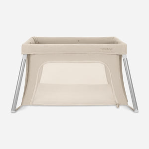 The Newton Baby Compact Travel Crib & Play Yard, shown on a white background, is beige with mesh sides, angled metal legs, a breathable mattress, a rectangular shape, and a zippered front opening.