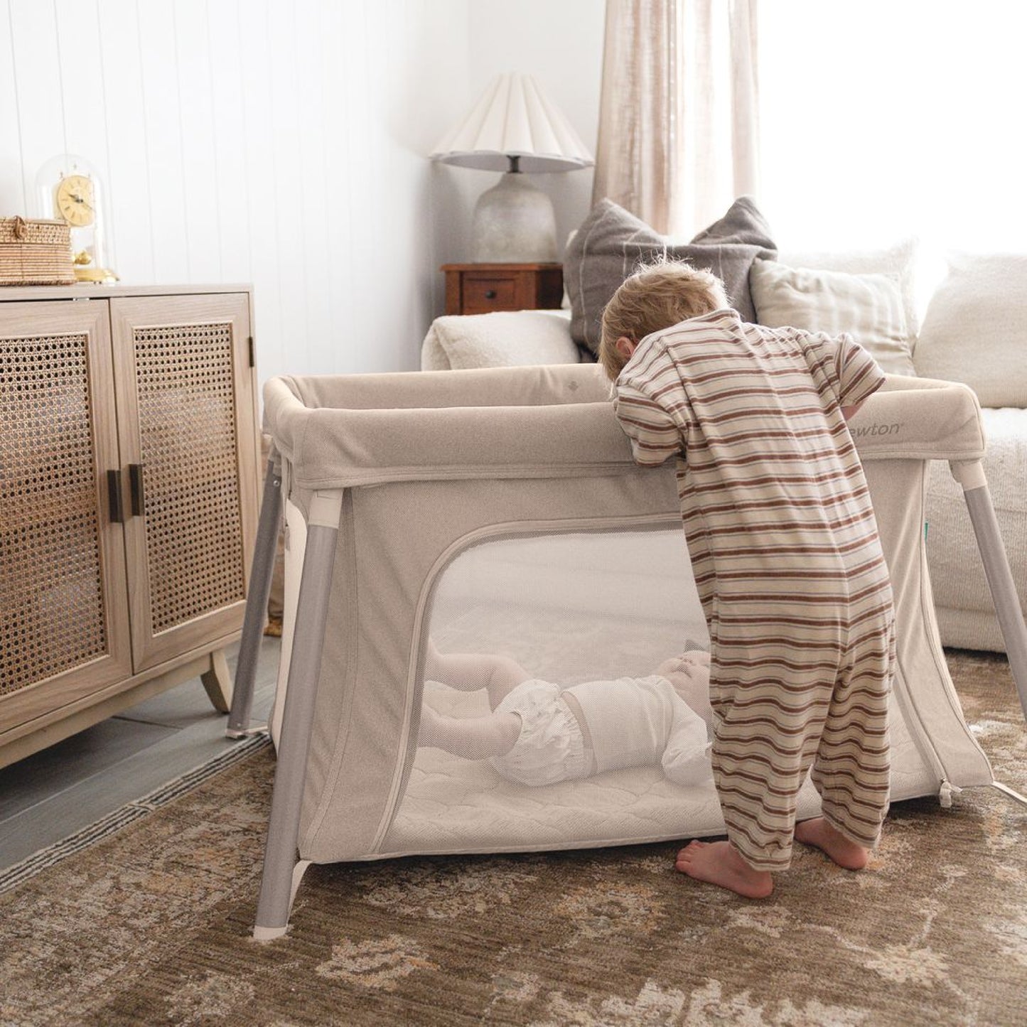 A toddler in striped pajamas peers into a Newton Baby Compact Travel Crib & Play Yard with a breathable mattress, where a baby lies inside, set in a cozy living room with soft lighting, a rug, and comfy furniture.