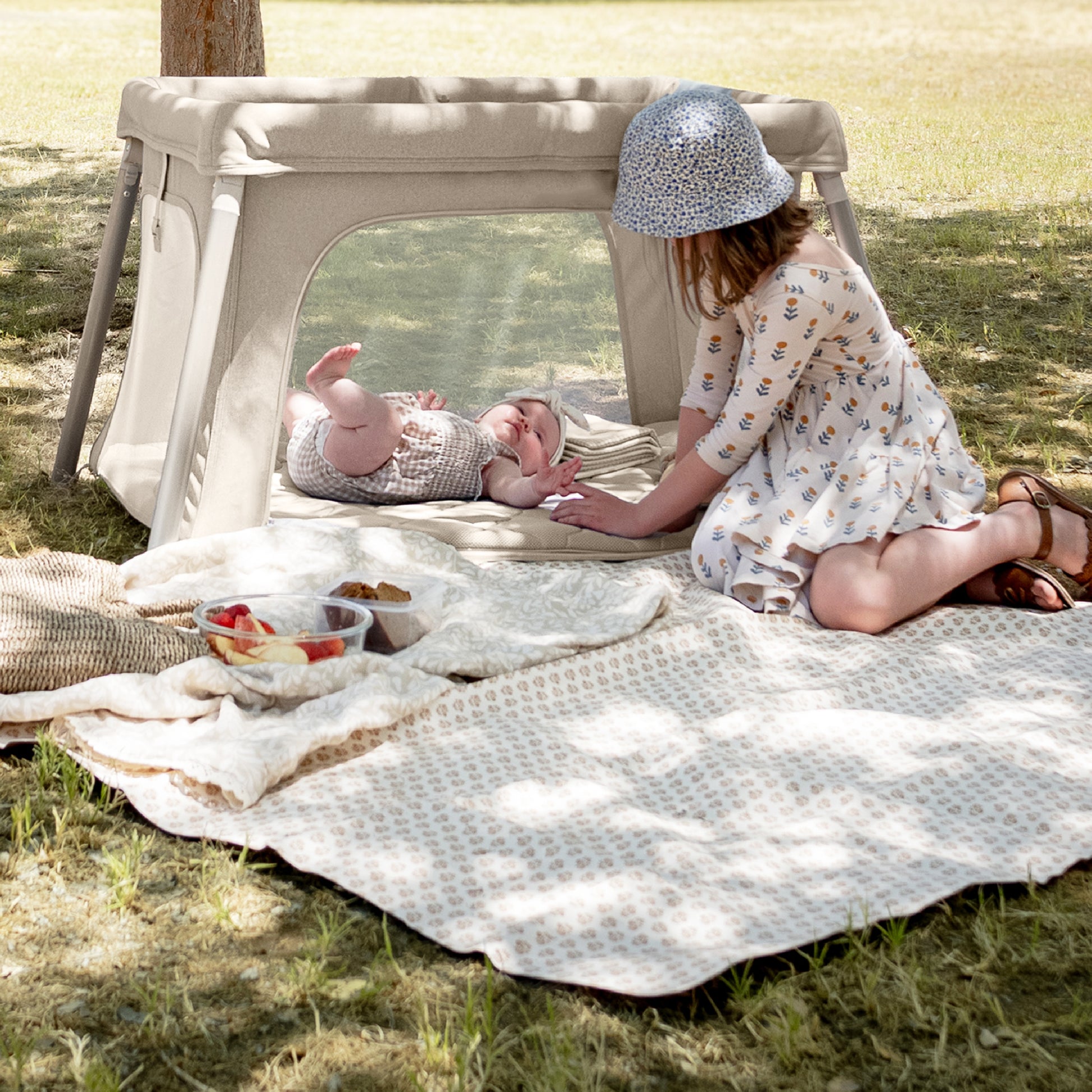 A young girl in a dress and sun hat sits on a blanket outdoors, gently touching a baby resting in the Newton Baby Compact Travel Crib & Play Yard, which features a breathable mattress. Picnic food and another straw hat are nearby under the tree’s shade. 999-5101-SAN