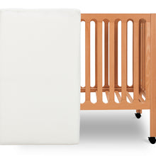 ALT text: "Newton Baby crib mattress placed vertically beside a light wooden crib with wheels on a white background."