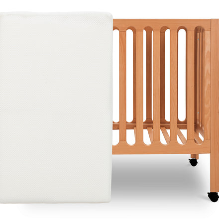 ALT text: "Newton Baby crib mattress placed vertically beside a light wooden crib with wheels on a white  background." skip