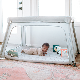 A baby lies on their tummy inside a soft, light gray playpen with a quilted mat, surrounded by soft toys including a green car and colorful blocks. Sunlight filters through windows in a bright, cozy room.