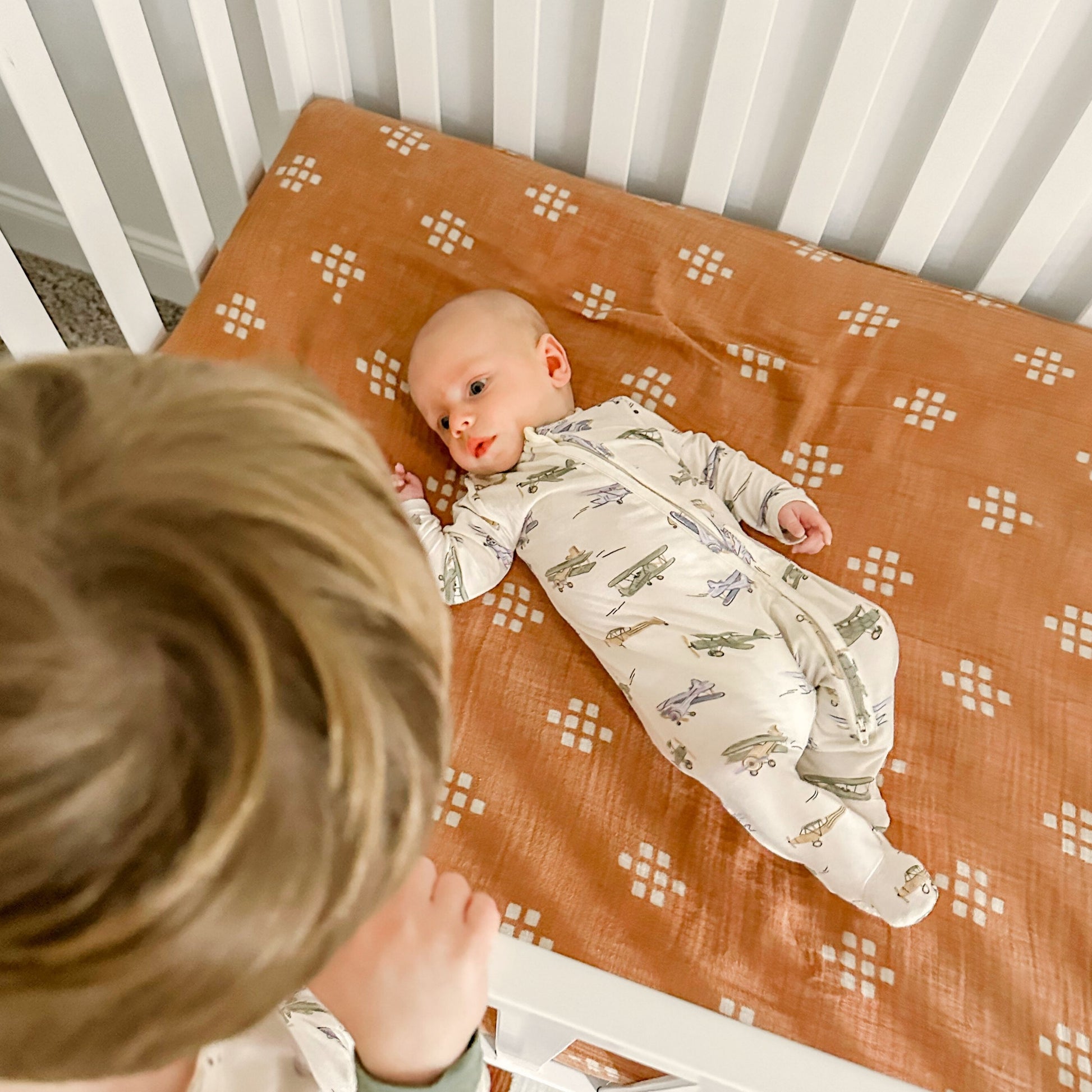 A baby in a printed onesie lies on a rust-colored crib sheet with white patterns atop a Newton crib mattress, while an older child looks down from the side of the dadada baby Austin Convertible Crib.